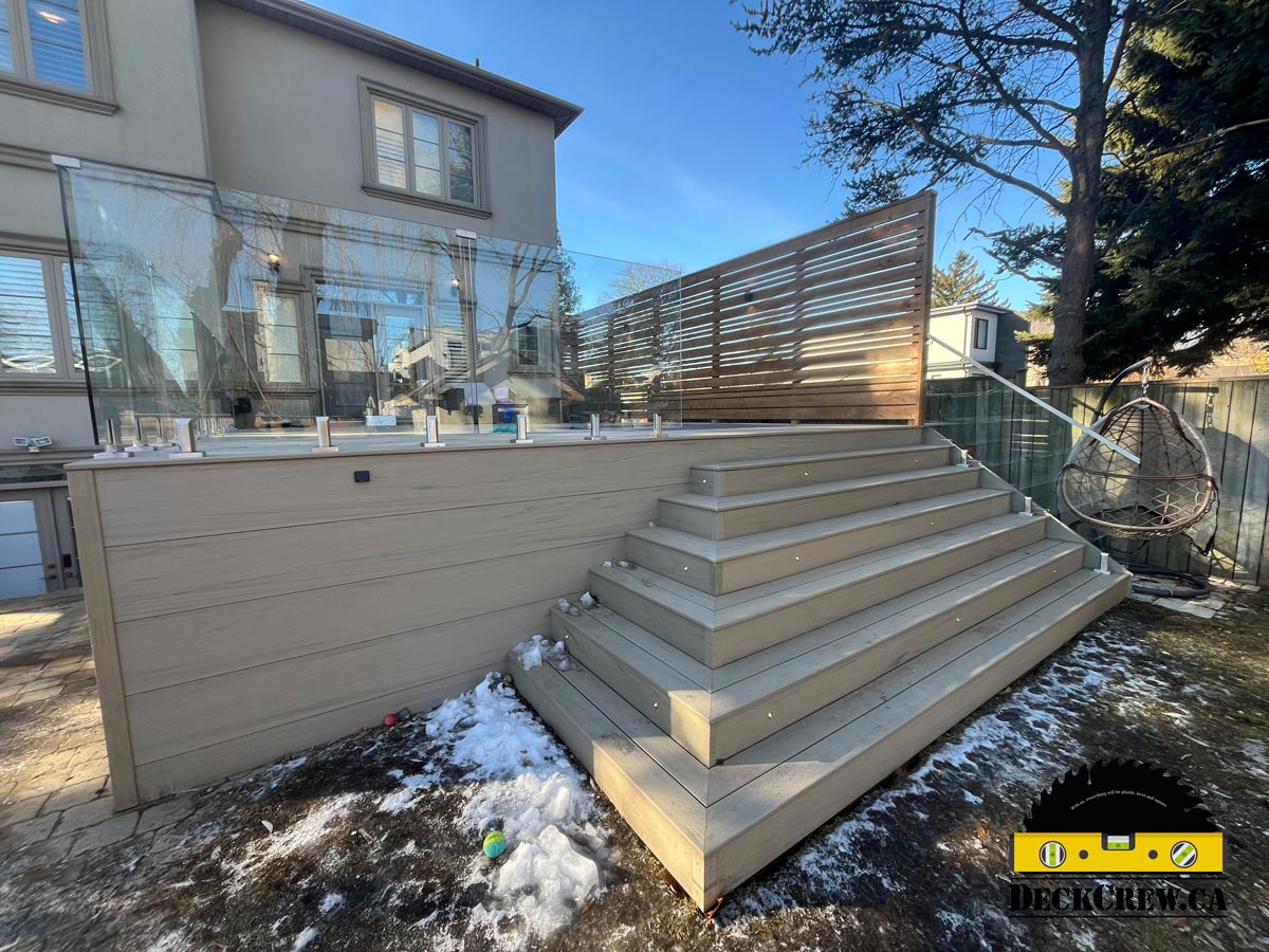 Composite deck with wide stairs, integrated step lighting, frameless glass railings, and privacy screen in a residential backyard.